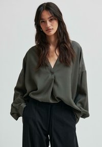 Blusa verde oliva con bottoni, scollatura a V, vestibilità ampia e maniche lunghe, abbinata a pantaloni neri a vita alta. Tessuto dalla texture liscia.
