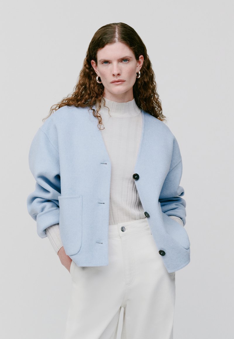 Lichtblauwe wollen cardigan met verlaagde schouders, twee zakken en zwarte knopen. Gestyled over een crèmekleurige geribde coltrui en witte broek.