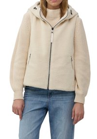 Frau in einer hellbeigen Fleece-Weste mit Kapuze, die über einem beigen Strickpullover und blauen Jeans steht, mit entspannten Händen an den Seiten.