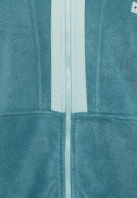 Teal fleecejakke med hvit glidelås og vertikale hvite striper. Myk tekstur, med glidelås foran og detaljerte lommer.
