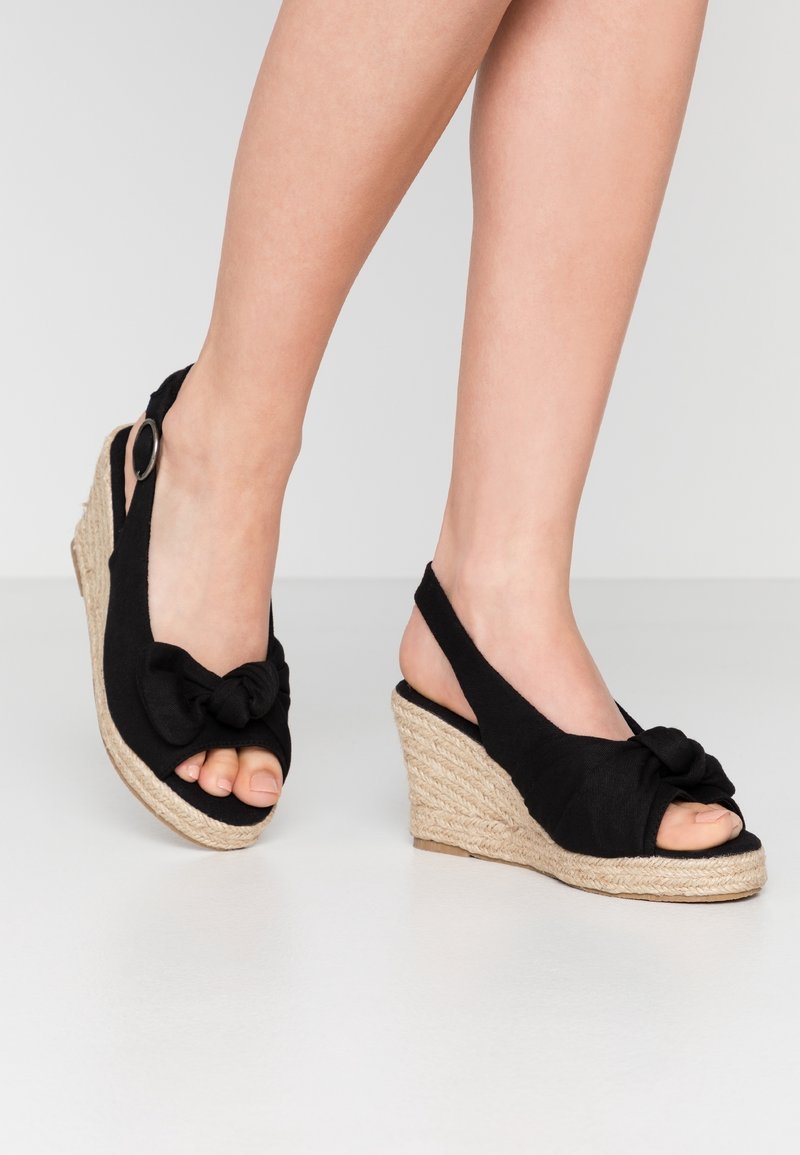 Jambes portant des sandales compensées en tissu noir avec orteils ouverts, détail noué à l'avant et talons compensés en jute tressée sur un sol blanc.