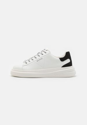 Biały niski sneaker z czarnym detalem na pięcie, z logo "GUESS" z boku, oczkami na sznurówki i grubą podeszwą na jednolitym tle.