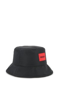 Chapeau seau noir en tissu lisse, avec un patch logo rectangulaire rouge portant le texte "HUGO" sur le côté. Large bord doux.