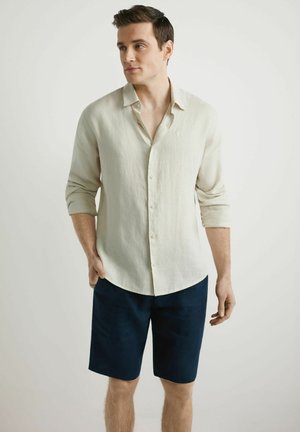 Mężczyzna ubrany w beżową koszulę na guziki z podwiniętymi rękawami i ciemnoniebieskie szorty, stoi z jedną ręką w kieszeni, patrząc na bok.