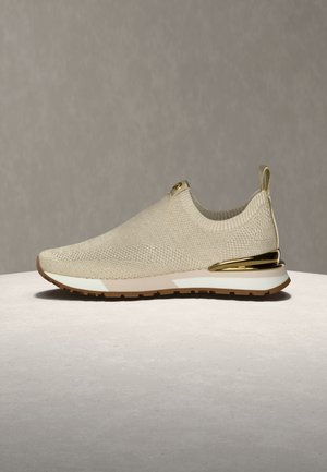 Beige knit sneaker with a cushioned sole, gold accents, and a loop at the heel. Features a flexible, breathable design without laces.