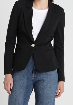 Mujer con blazer negro ajustado de un solo botón, camisa blanca y jeans azules, de pie frente a un fondo neutro.