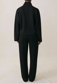 Czarna dopasowana marynarka i pasujące spodnie wykonane z miękkiej tkaniny. Marynarka ma wysoki kołnierz i opadające ramiona, a spodnie są proste.