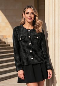 Czarno-biała marynarka w kratkę z srebrnymi guzikami, dwoma przednimi kieszeniami i długimi rękawami, noszona na czarnej spódnicy. Strukturalna faktura materiału.