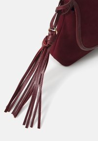 Sac à main en suede bordeaux avec une sangle en cuir tressé et de longs accents en frange. Bords lisses et texture douce avec une forme arrondie.