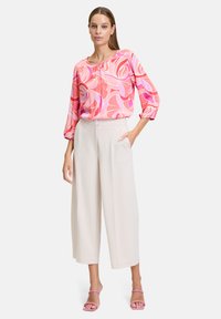 Blusa rosa con motivo floreale e maniche larghe, abbinata a pantaloni beige a vita alta. Il modello indossa sandali rosa con cinturini.