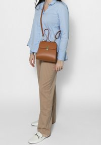 Borse a spalla in pelle marrone con forma rettangolare, accessori in oro e chiusura a pattina. Indossata in modo casual con pantaloni beige a righe.