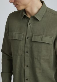 Camicia verde oliva con bottoni, realizzata in tessuto testurizzato, caratterizzata da due tasche sul petto con pattine e bottoni neri lungo la parte anteriore.