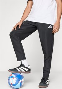 Μαύρο αθλητικό παντελόνι με στενά πόδια, λευκό λογότυπο με τρεις ρίγες, σε συνδυασμό με μαύρα και άσπρα αθλητικά παπούτσια και πολύχρωμη ποδοσφαιρική μπάλα κάτω από το πόδι.