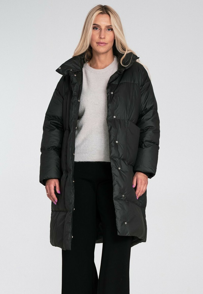 Czarna kurtka puchowa z wysokim kołnierzem, zapięciem na napy i pikowanym wzorem. Nosi się ją na jasnoszary sweter i czarne spodnie. Gładka faktura.