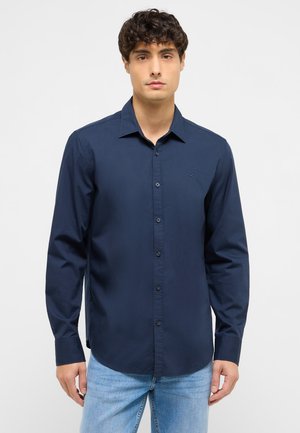 Mann trägt ein dunkelblaues, langärmeliges Hemd mit Knöpfen und hellblaue Jeans, blickt nach vorne vor einem schlichten Hintergrund.