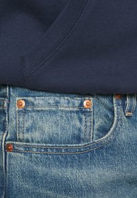 Zbliżenie na kieszeń niebieskich dżinsów z mosiężnymi nitami, częściowo zakrytą brzegiem granatowego swetra z dzianiny.