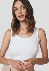 Biała bawełniana bluzka na ramiączkach z okrągłym dekoltem i szerokimi ramiączkami. Modelka nosi złoty pierścień, ma gładką skórę i falujące, średniej długości brązowe włosy.