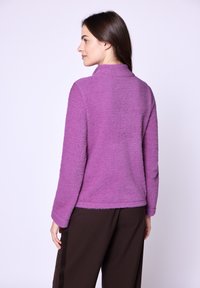 Maglione in pile viola con colletto alto e maniche lunghe, caratterizzato da una finitura testurizzata. Abbinato a pantaloni marroni a righe sottili.
