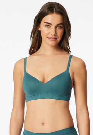 Woman with long brown hair wearing a teal wireless bra with adjustable straps, facing forward against a white background.