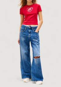 T-shirt rossa aderente con logo a cuore bianco abbinata a jeans denim blu larghi con un grande taglio sul ginocchio. Sneakers bianche semplici.