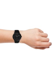 Montre noire avec un cadran rond, design minimaliste, bracelet en silicone et marquages simples sur le cadran, reposant sur une main.
