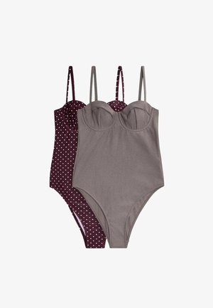 Jeune femme aux cheveux courts et foncés portant un maillot de bain une pièce gris, debout devant un fond blanc uni, regardant légèrement sur le côté.