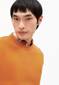 Giovane uomo con capelli neri corti che indossa un maglione arancione sopra una camicia a quadri, con uno sguardo leggermente di lato su uno sfondo bianco.