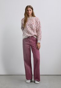 Frau steht und trägt einen rosa und beige Leopardenmuster-Pullover, lila Zebra-Muster weit geschnittene Hose und weiße Sneakers vor einem schlichten Hintergrund.