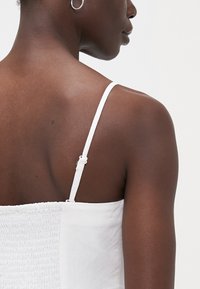 Valkoinen, hihaton paita, jossa on ohuet olkaimet ja smockattu pinta takana. Paita on minimalistinen designiltaan ja sen kangas on sileä.