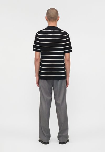 Άνδρας με ξυρισμένο κεφάλι, που φοράει μαύρο-λευκό κοντομάνικο πουκάμισο με οριζόντιες ρίγες και γκρι φαρδιά παντελόνια, στέκεται γυρισμένος με την πλάτη.