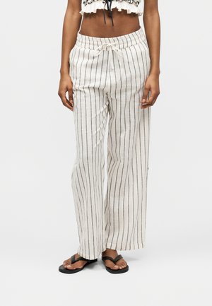 Kobieta nosząca luźne białe spodnie z czarnymi pionowymi paskami i sznurkiem w pasie, połączone z czarnymi klapkami.