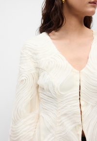 Krémová blúzka s textúrovanou, vlnitou tkaninou, hlbokým V-výstrihom a gombíkovými uzávermi na prednej strane. Dlhé rukávy s jemným splývaním.