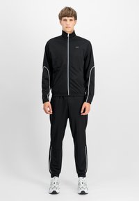 Juoda sportinė striukė su aukštu apykakle ir baltomis siūlėmis, derinama su juodomis sportinėmis kelnėmis. Modelis vilki sidabrinius sportinius batus.