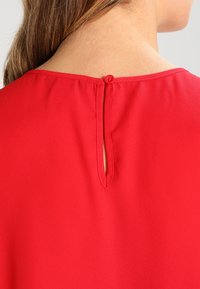 Rød bluse med et nøglehul design på bagsiden, knaplukning og glat stofstruktur. Udhullet er rundt og uden sømme.