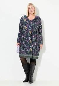 Mörk långärmad klänning med paisleymönster i rosa, grön och blå, med en insydd midja och dekorativ fåll. Svarta cowboystövlar.