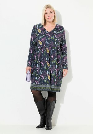 Langärmliges Kleid aus dunklem Stoff mit Paisleymuster in Pink, Grün und Blau, mit geraffter Taille und dekorativem Saum. Schwarze Cowboy-Stiefel.