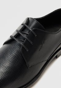 Chaussure noire en cuir à lacets avec un bout texturé et un empeigne lisse, dotée de lacets gris et de détails de couture subtils.