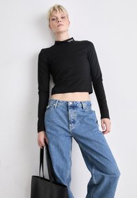 Haut noir à manches longues et cropped avec un col haut et un petit logo. Assorti avec un jean en denim bleu clair, taille haute et ample, et un sac fourre-tout noir.