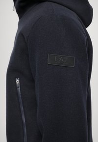 Námornícky modrá fleece mikina s hladkým povrchom, s bočnou zipsovou kapsou a čiernou koženou nášivkou na pleci.