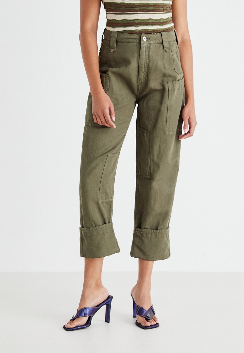 Pantalones cargo verdes de corte relajado, con grandes bolsillos laterales y puños enrollados. Combinados con sandalias de tacón moradas.