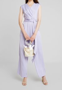 Lavendel jumpsuit med omslagsdesign, korta ärmar och knytmidja. Matchad med en genomskinlig handväska med en krämfärgad dragsko-påse.