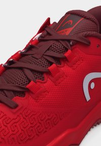 Nahaufnahme eines roten Sportschuhs mit dunkelroten Schnürsenkeln, strukturiertem Mesh-Zungenbereich und erhabenem Muster an der Seite nahe dem weißen Logo.