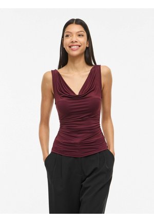 Uśmiechnięta kobieta w bezrękawnym bordowym marszczonym topie i czarnych spodniach, stojąca z rękami w kieszeniach na białym tle.