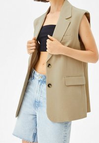 Beżowy bezrękawnik z wykładanym kołnierzem, dwoma guzikami i przednią kieszenią, zestawiony z czarnym topem na krótko i jasnoniebieskimi szortami dżinsowymi.