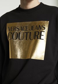 Černá mikina s lesklým zlatým obdélníkovým motivem s nápisem "VERSACE JEANS COUTURE" vytištěným černými tučnými písmeny.