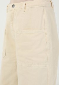 Pantaloni in cotone beige con una vestibilità rilassata, dotati di una tasca frontale, chiusura con bottone metallico e dettagli di cucitura in vista.