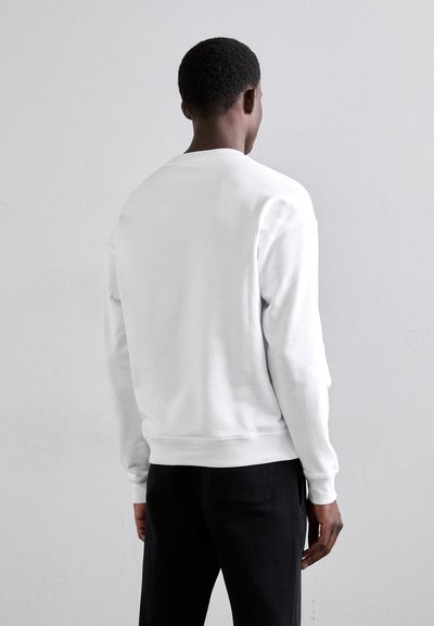 Sudadera blanca con cuello redondo, puños y dobladillo acanalados. Textura suave, diseño ajustado, y llevada sobre pantalones negros. Se muestra vista trasera.