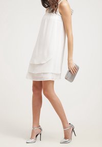 Vit lagerklänning med ett flödande design, parat med silverhöga klackskor och en grå handväska. Slät, lätt tygtextur.