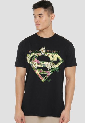Young man wearing a black t-shirt with a floral Superman logo and the text "My Mom My Hero" looking to the side on a plain background.