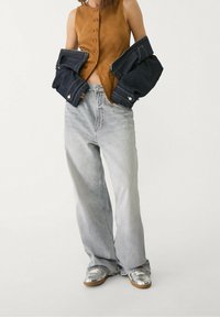 Bruin suède mouwloos topje met knoopdetail, gecombineerd met lichtgrijze wijde jeans en zilveren sneakers. Spijkerjack over de arm gedrapeerd.